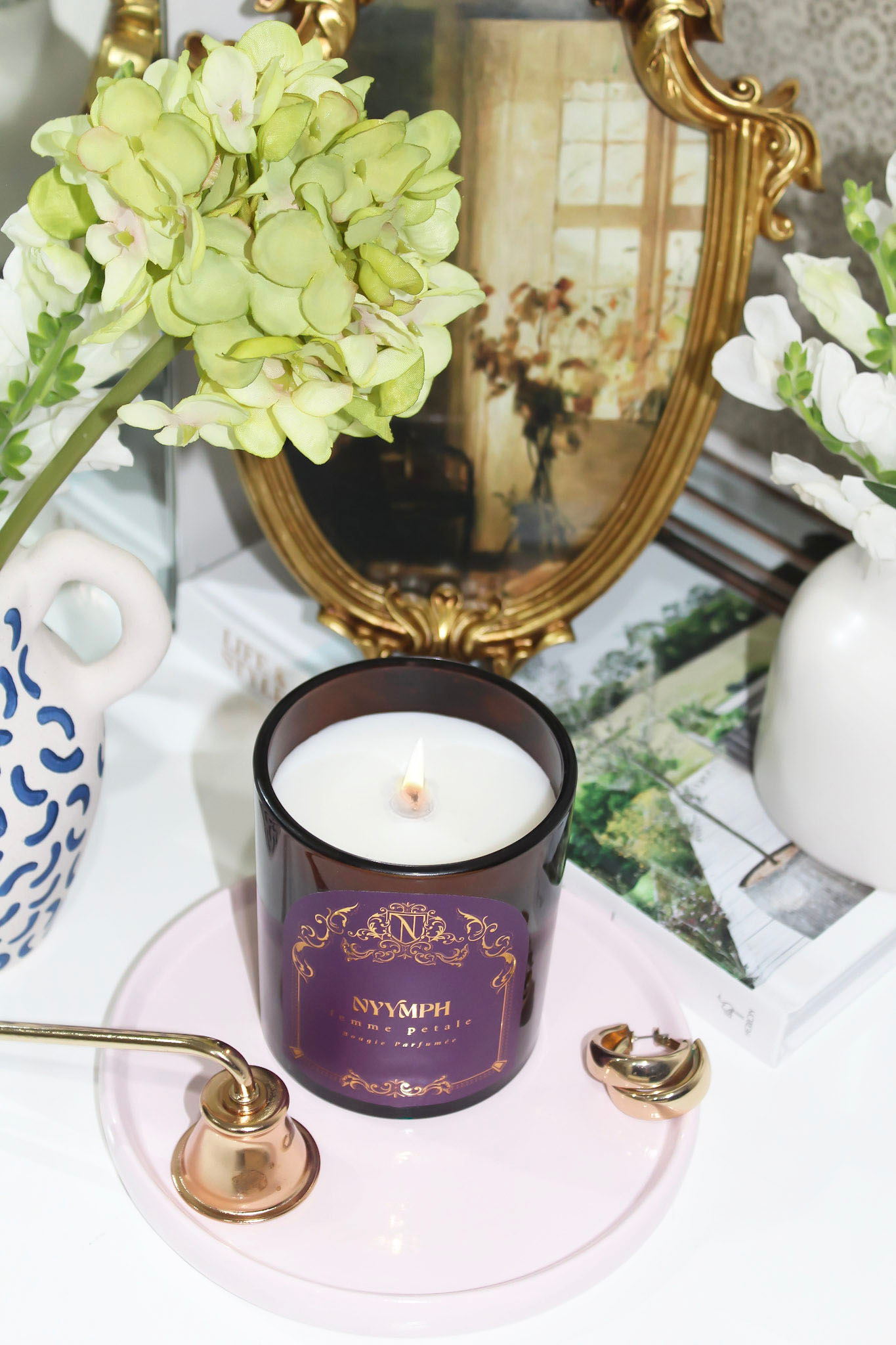 NYYMPH Femme Petale lit candle on pastel pink tray, styled with gold snuffer, floral stems, and vintage mirror decor.
