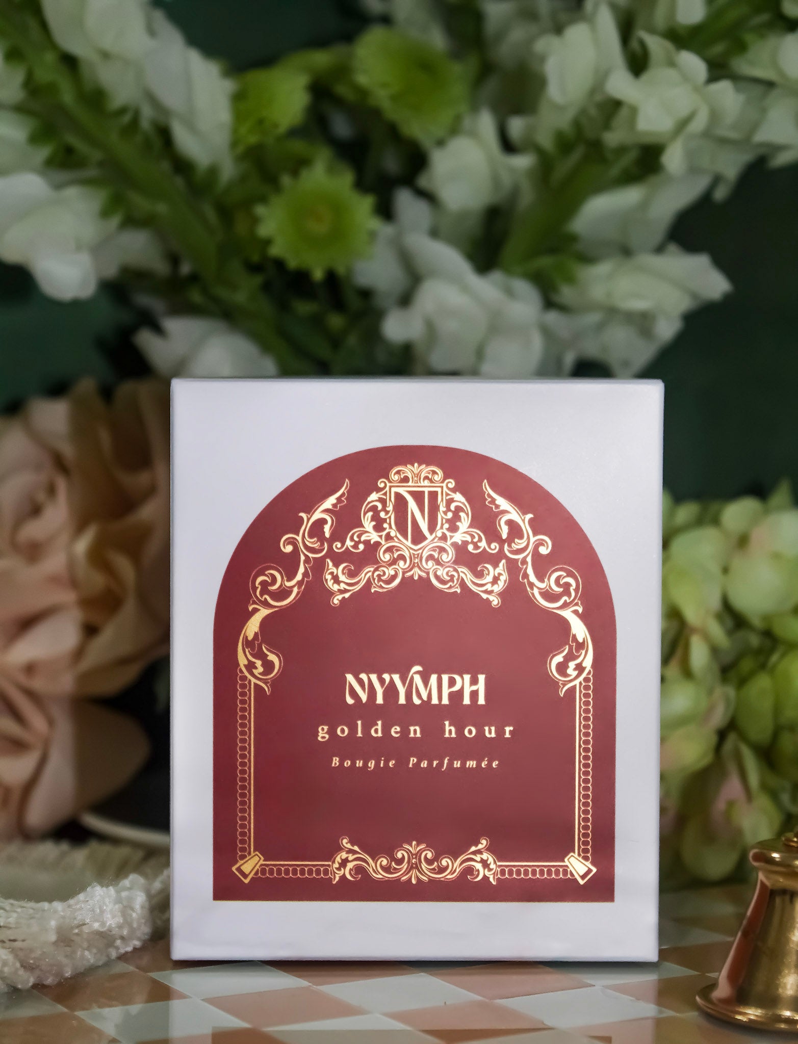 NYYMPH Golden Hour candle packaging – luxury box with gold and burgundy design, styled with soft floral arrangement.