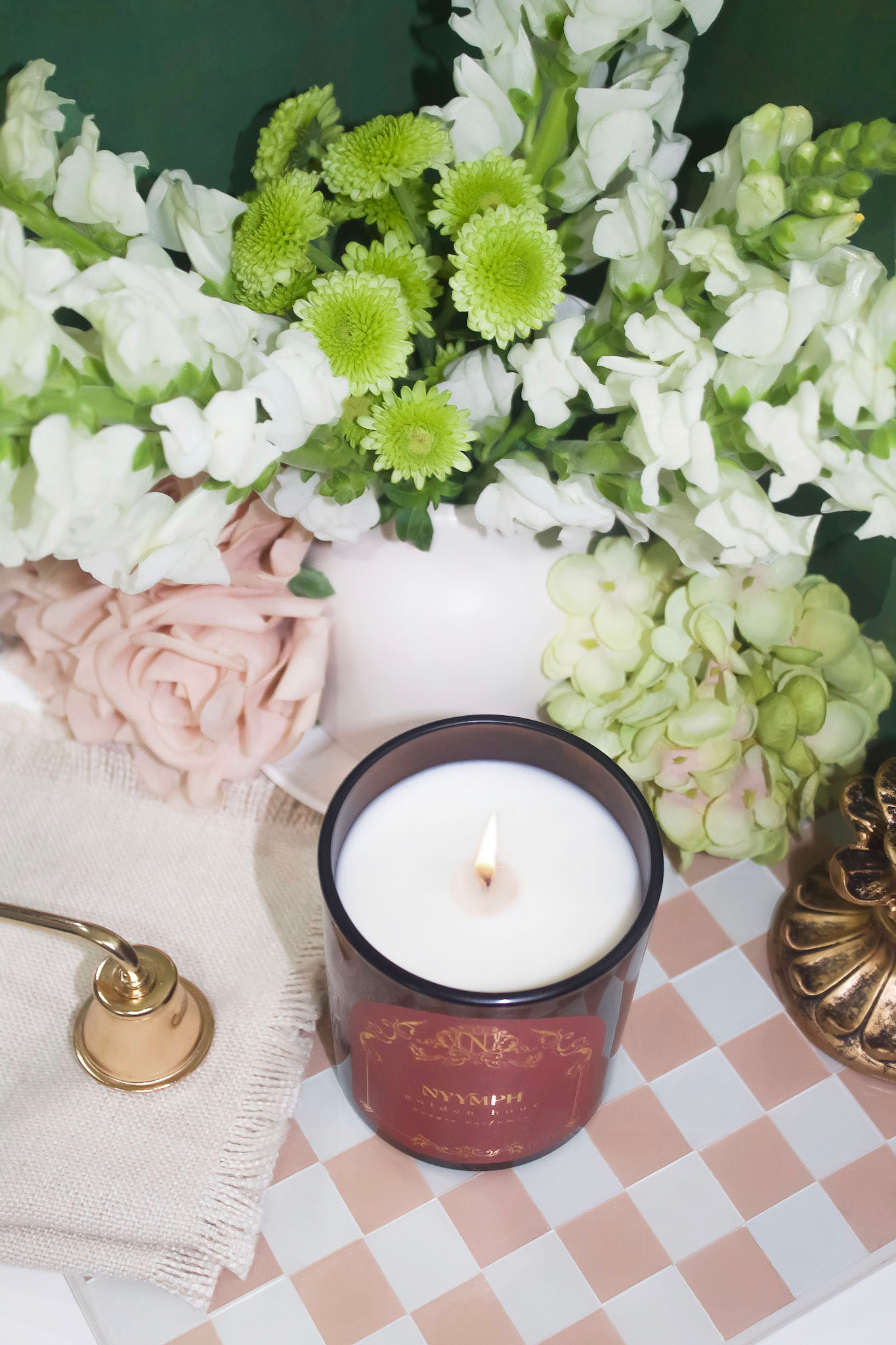 NYYMPH Golden Hour candle lit – soy-coconut wax in maroon vessel styled with floral decor, brass snuffer, and checkered cloth.