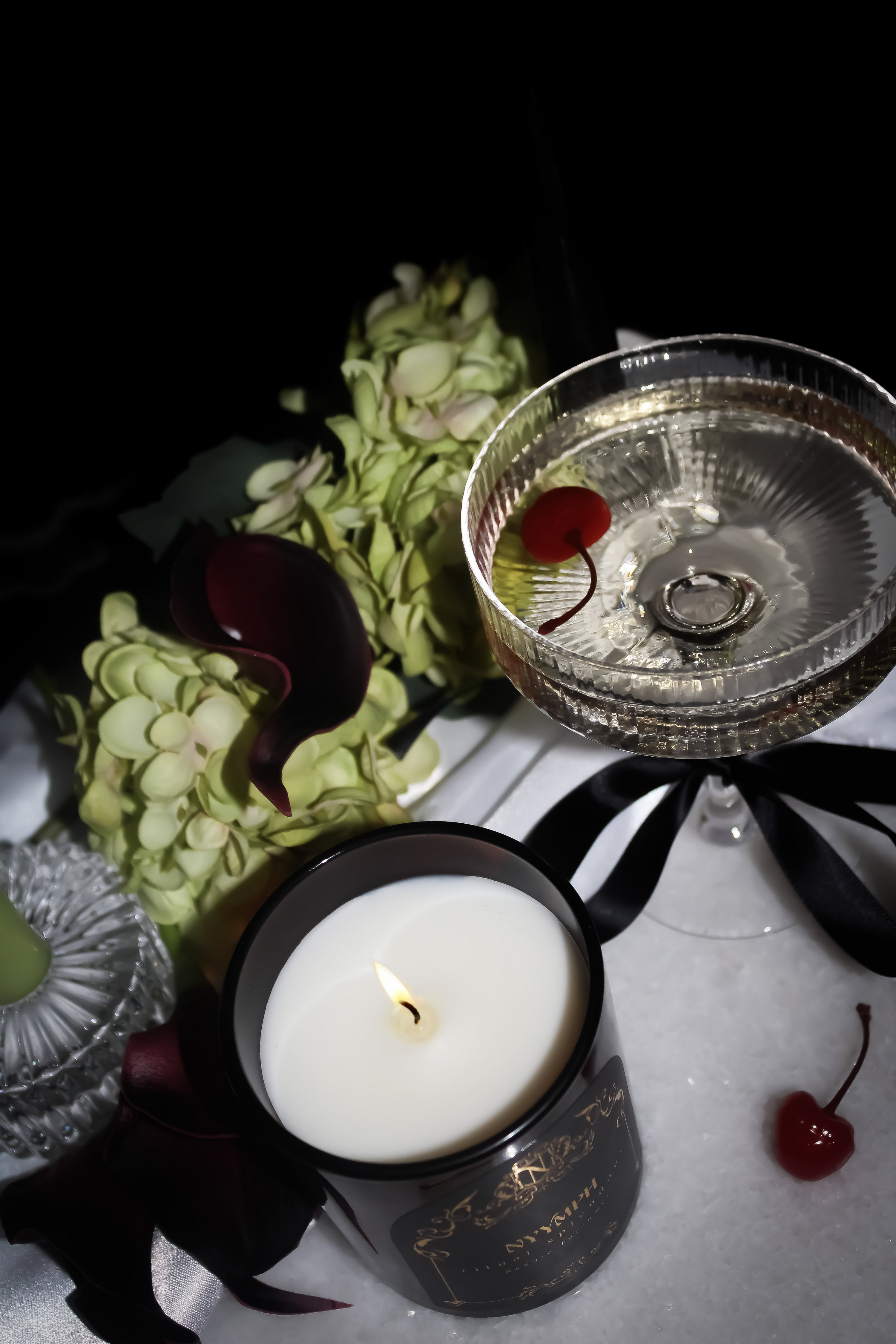 NYYMPH Ritual Splendour candle lit beside cocktail glass with cherry, hydrangeas, black ribbon and rose petals – styled in a moody luxury setting.