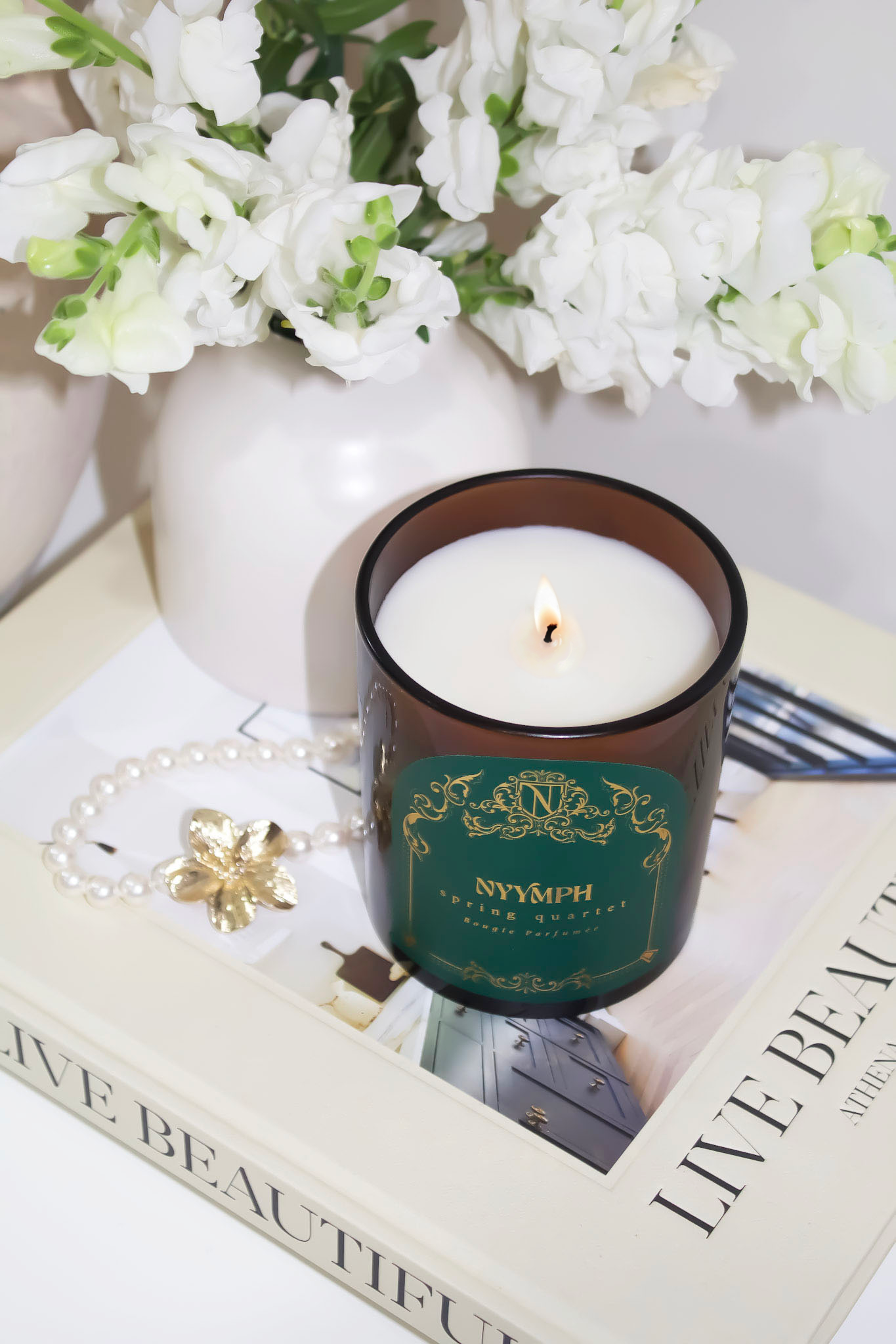 Lit NYYMPH Spring Quartet candle styled with white blooms, pearl bracelet, and coffee table book for a chic, serene decor scene.