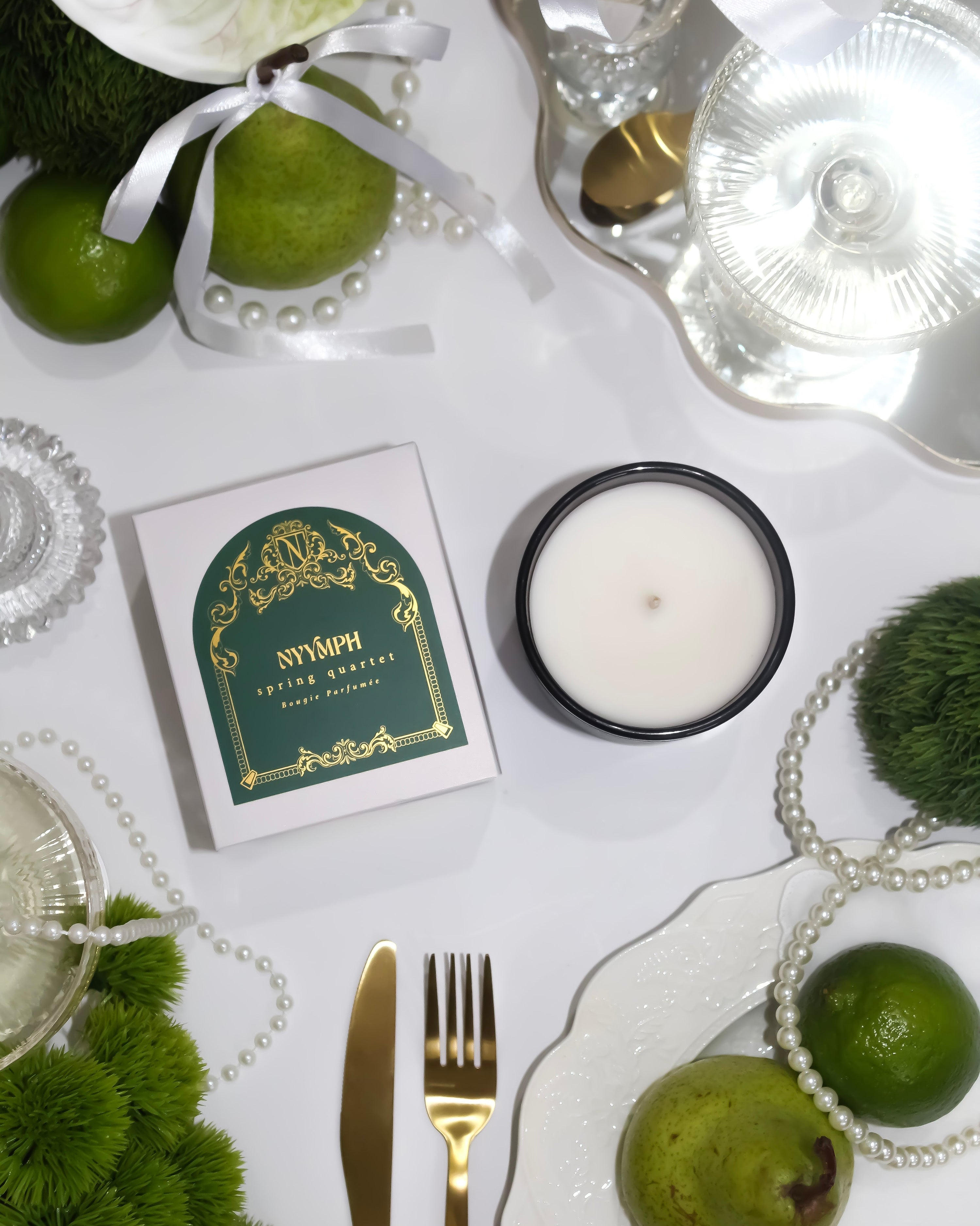 NYYMPH Spring Quartet candle with green label and box, styled on a white table with limes, ribbon, and pearls – a lush spring tablescape.